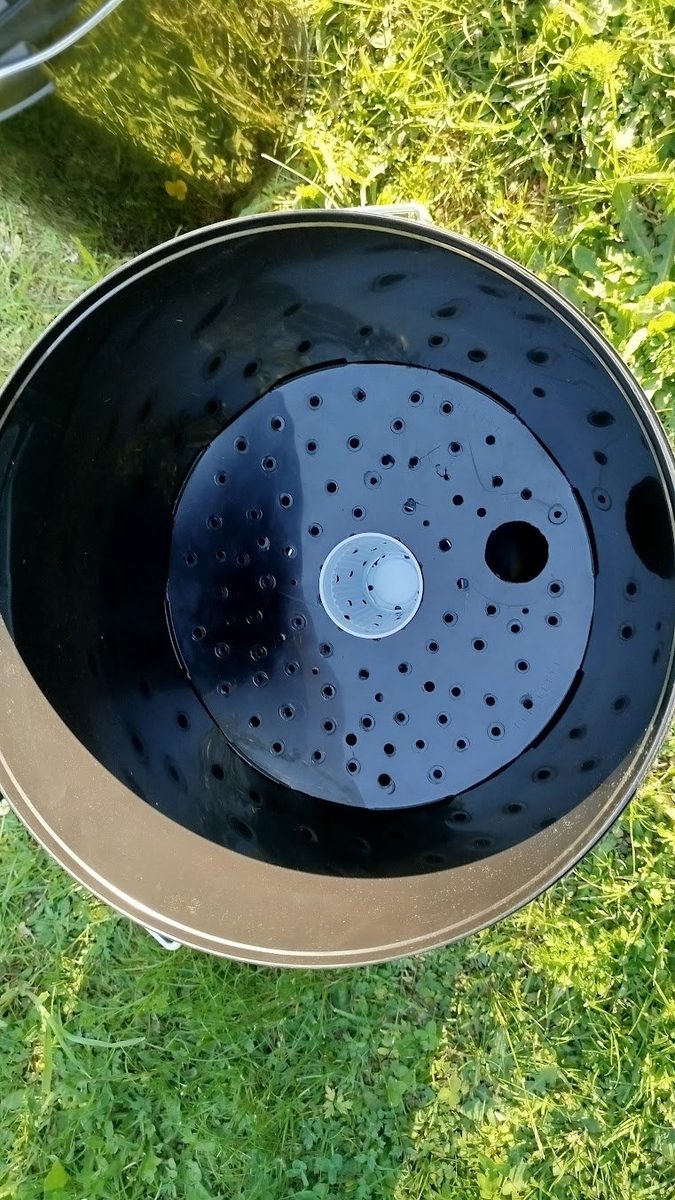 Draufsicht in einen schwarzen Eimer: selbstgebauter Deckel-Erdrost mit perforiertem weißen Wickdocht-Becher in der Mitte, Belüftungslöcher rundum, größere Öffnung für Gießrohr.
